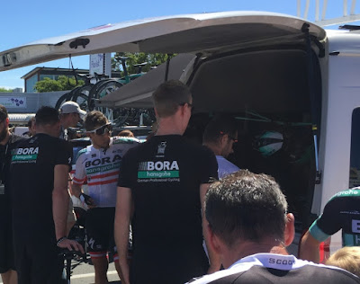 At the back of the open bus, Lukas Pöstlberger stands amongst a group. He is wearing a white jersey with the Austrian flag colours of red and white emblazoned on it.He is looking into the back of the bus with his white framed sunglasses raised onto his forehead.