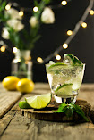 A clear sparkling liquid in a glass and a lime on a wooden table with lemon, lights and a clear vase with flowers in the background.