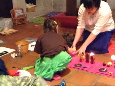 This is a rather jumbled picture - there are several baskets visible on the left, as well as a spray bottle of cleaner and a crumpled paper towel, and some papers and crayons. The focus of the picture, however, is of the Storyteller in the back right of the photo, kneeling, and holding the edges of a candle holder, and a young girl in the center of the photo who is down on one knee and leaning forward to snuff out the candle with a small snuffer.