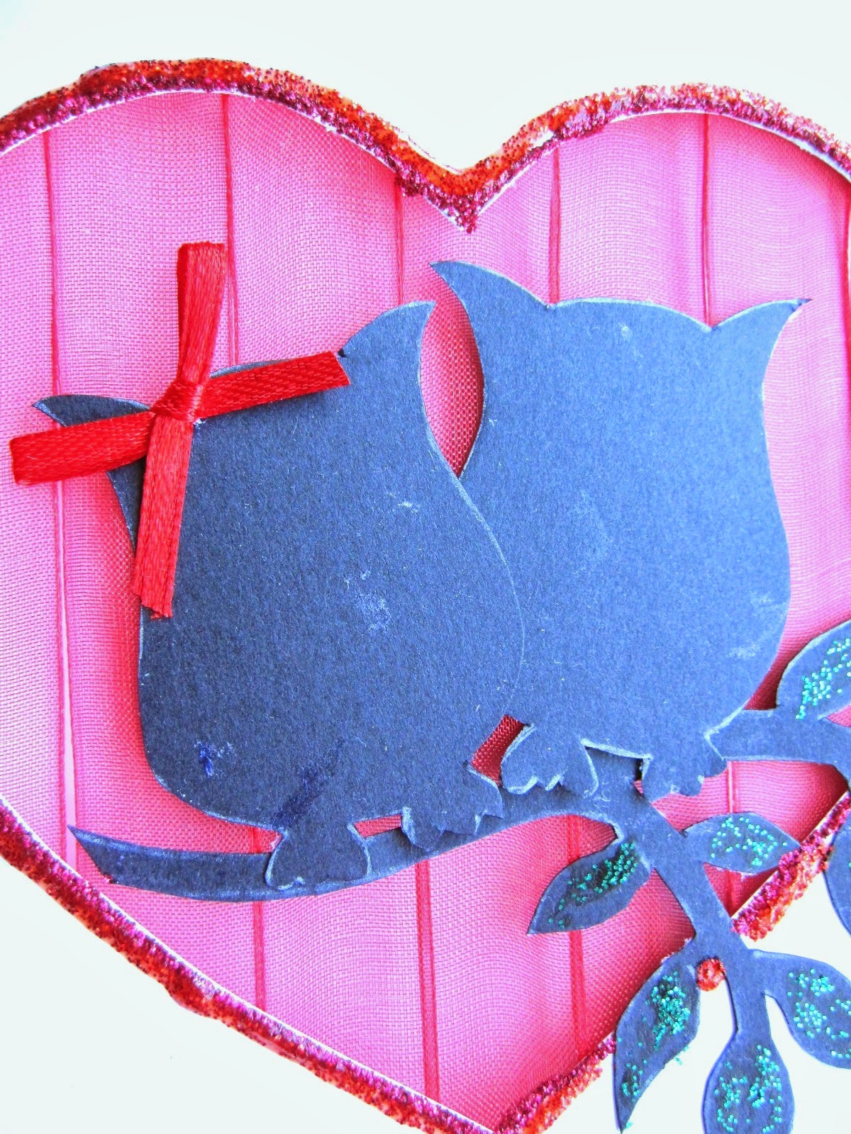 detalle de tarjeta blanca de felicitación con corazón calado con fondo de gasa roja y siluetas delante de dos búhos y rama de árbol con detalles decorados con glitter glue rojo y verde