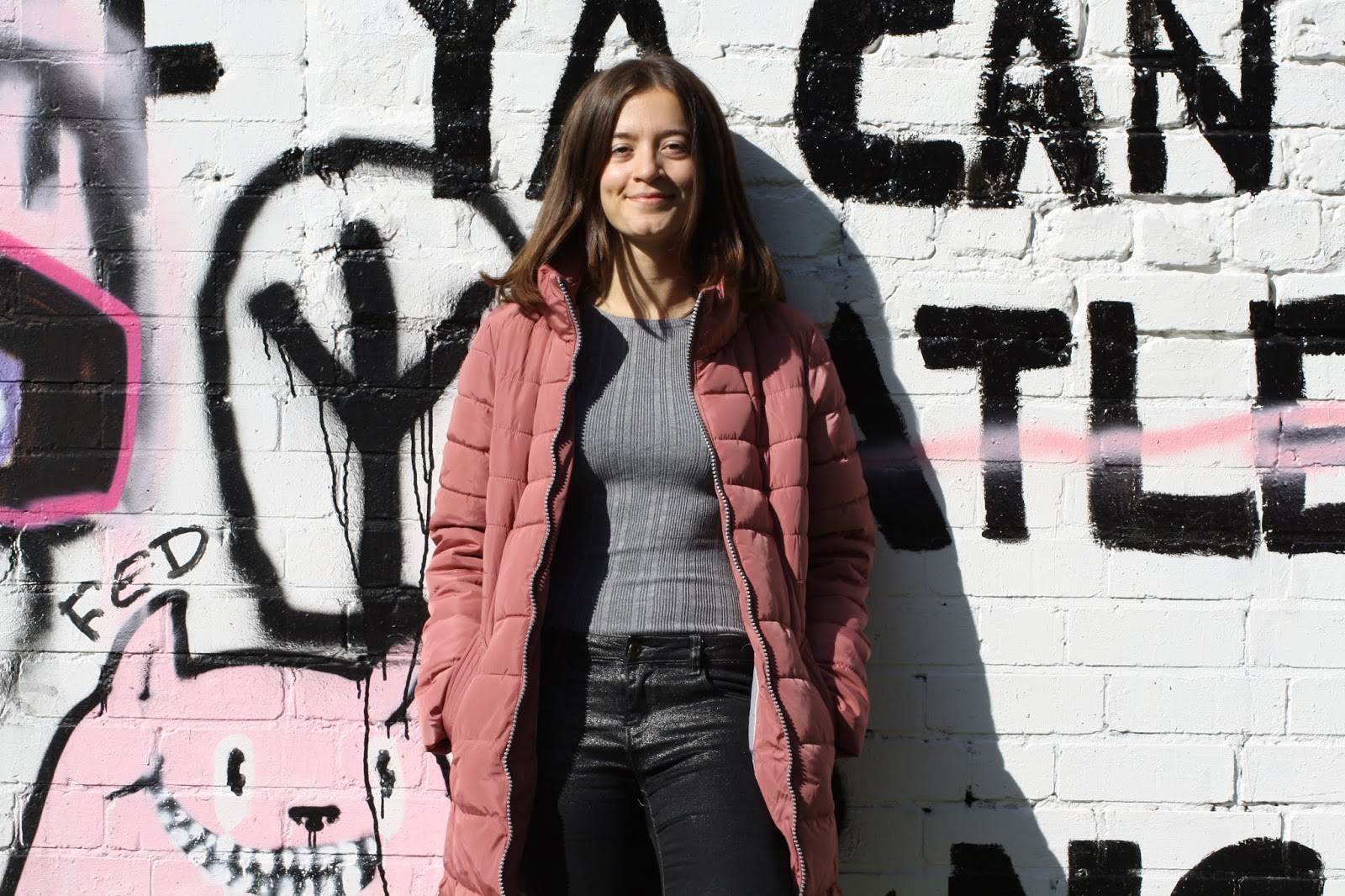 Abbey, leaning against a graffiti wall, smiles at the camera, hands in pockets