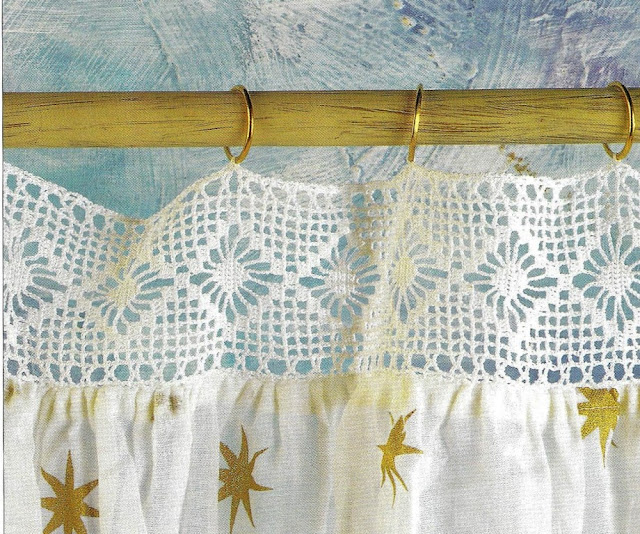 From a light wooden curtain rod hangs a fabric curtain with a heading made of white filet crochet with a spider stitch frieze.  The curtain fabric is white with gold stars.  The heading hangs from the rod by narrow gold or brass rings which have been threaded through the top edge of the filet crochet.