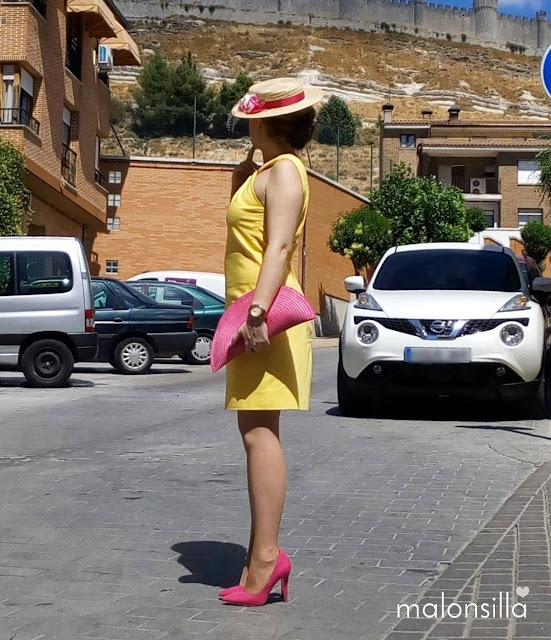 Invitada boda con vestido amarillo corto con escote en la espalda, canotier de flores de copa baja en fucsia y bolso abanico fucsia 