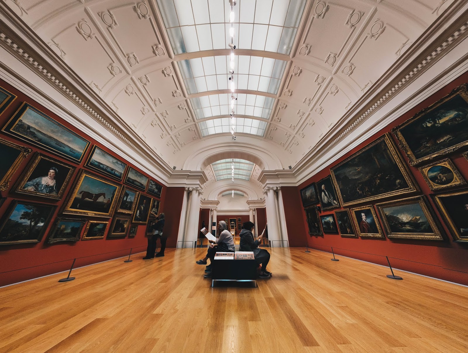 A photo looking down a room in an art gallery with paintings on the walls on either side, with a bench in the middle with people sitting and sketching, and two people looking at the paintings on the left - photo taken in Auckland Art Gallery Toi o Tāmaki, Auckland, New Zealand