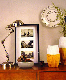 Catalogue ad showing a silver lamp, a bowl of pinecones, a three-section picture, two wooden vases (dip-painted white, one with a sprig of bush), and a decorator plate on the wall.