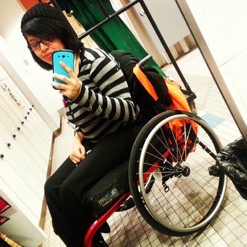 Photo of an Asian young woman wearing glasses, a black and white striped shirt, sitting in a wheelchair, taking selfie with a smartphone