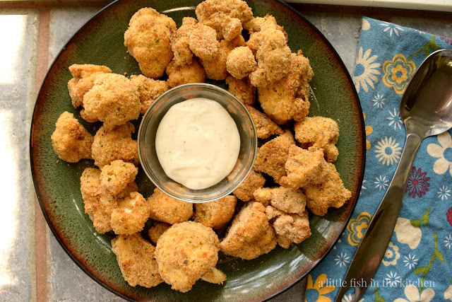Crispy oven-fried cauliflower is ready to serve on a dark green ceramic plate. A small clear, glass bowl of ranch dressing sits in the center of the breaded cauliflower for dipping and sharing. A blue linen tea towel with a bright floral print is folded beside the plate with a metal serving spoon on top. 