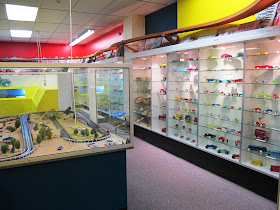 Rows of metal cars displayed on shelves in glass cabinets lining the walls. In the front left is a slot-car set up under perspex.