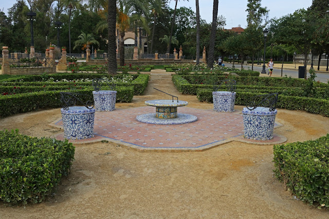 Avenida interior de un parque con un monumento circular con un reloj de sol y un camino de setos entre palmeras.