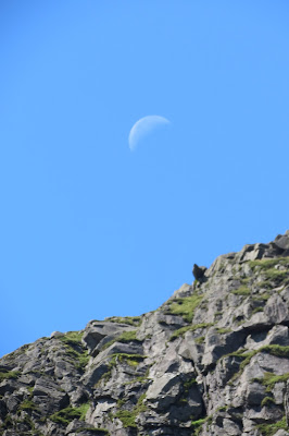 A rock face with a dark shadow near its edge. The moon sits above in a cloudless blue sky.