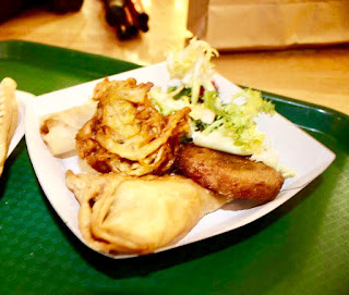 A selection of lightly brown Indian treats in the shape of circular patties and slightly oval lightly brown battered ovals with a light green salad consisting of light green shredded lettuce and dark green cubed pieces of Cucumber on a white square polystyrene plate on a light wooden table on a light background. 