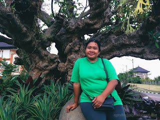 Woman Sitting On The Stone Under A Beautiful Ornamental Tree In The Garden At Badung, Bali, Indonesia