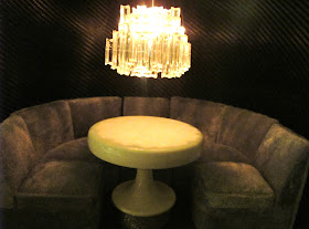 Modern one-twelfth scale miniature nightclub scene of a curved booth is grey velvet, with a round table in the middle and a large crystal chandelier hanging above it. 