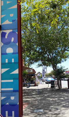 Along the left edge is a vertical banner which reads "ArtsCentre" in bold, blue, overlapping capital letters. Beyond the banner one can follow the footpath to rest on a wooden bench under a tree or continue to the cafe and shops. The verandah of a cafe can be seen next to the banner and under the below the tree canopy in the picture.