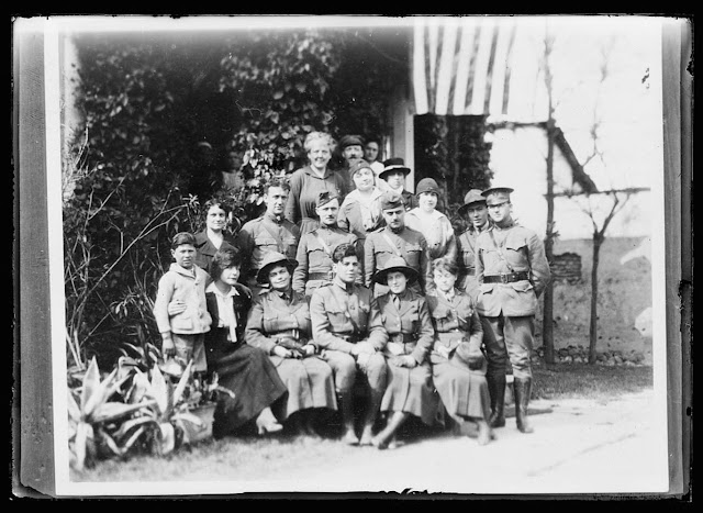 Red Cross personnel at Monastir. From left to right, back row, Miss Rogers and two Serb workers, next row, Miss Rose and Mrs. Freeman, next row: Serbian workers, Major Rogers Perkins, Captain Austin, Captain Pfotzer, Miss Crosley, Lieut., Paul Ivanichevitch, Lieut. Adams, front row, Serbian orphan, Miss Mountain, Dr. Keyes, Capt. Lanning McFarland, Dr. Flood, Miss Saxton. The two army officers in the group, Capts. Austin and Pfotzer organized the municipal health department of Monastir and are members of it. Drs. Keys and Flood, the two Red Cross women, have been the Balkans for a year and a half. During the Allied advance they worked in the front line dressing stations in the capacity of surgeons for days
