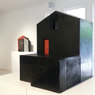 View of an art gallery exhibition with two Alex Asch assemblage art pieces in black and white, in the shape of buildings.