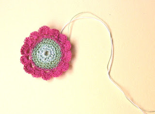A crocheted flower worked in fine size 80 thread. The 40 mm diameter flower has a 22 mm green centre disk, with 14 hot pink petals around it. Two threads can be seen to the right of the flower which will be used to attach the flower to a card.