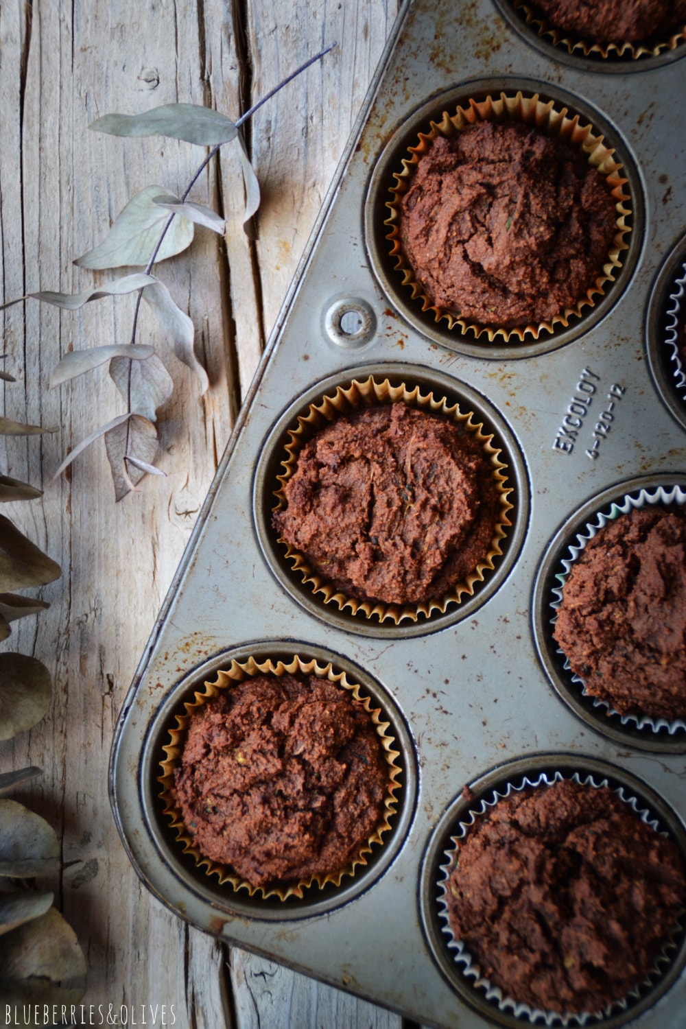 Cocoa muffins in old retro metallic pan, over old wood table, dark background, dry eucalyptus leaves, linen grey kitchen cloth
