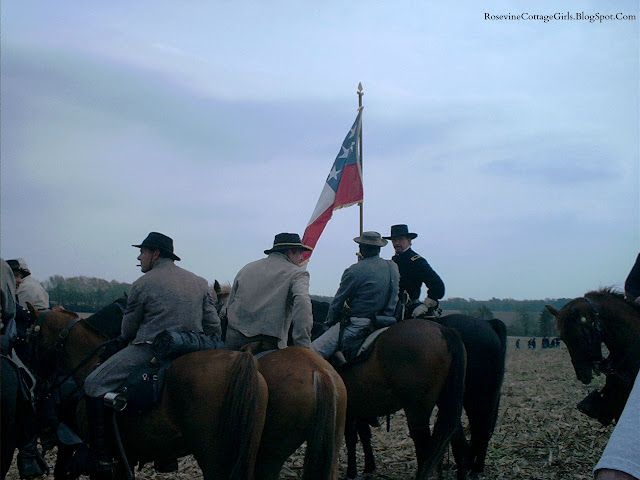Photo of an overcast day in November. Officers sit on horses awaiting orders to go into battle. Another officer on his horse holds aloft the flag. Behind them is a cornfield and soldiers on foot. This is the reenactment of the Battle of Spring Hill Tennessee an American Civil War Battle by RosevineCottageGirls.com