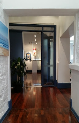 After entering the previous blue framed doorway, the view from the corridor shows polished wooden floors, whitewashed walls with artwork displayed. On the left wall one can see a painting of a shoreline in blue and sand colours.  Straight ahead is a double doorway framed by windows in blue frames. The left door appears closed with a pot plant in front of it. The right doorway is open to reveal a denim jacket and a crocheted bikini on the far wall of the main gallery.