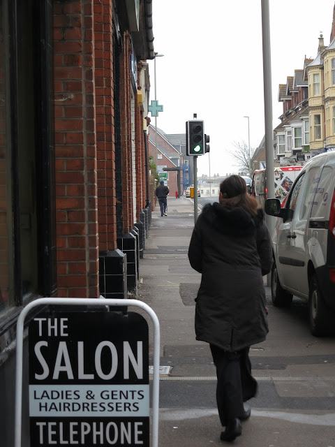 Woman walks down urban street past cars, lamposts and a hairdresser's sign.