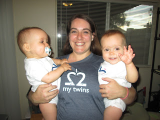 picture of mom holding baby twins, one pulling on her shirt the other waving at the camera