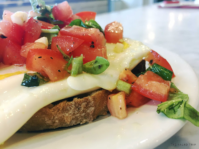 The Salad Trip: Savory Breakfast Bruschetta in the Kitchen A white plate on a white marble counter in a kitchen with bruschetta on trop of eggs and bread.