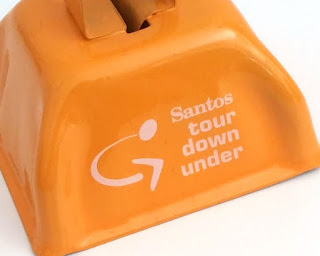 An orange pressed metal square cowbell with the Tour Down Under logo stamped on it in white. The words say "Santos tour down under"