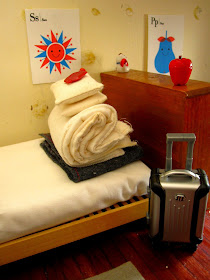 Miniature bed, stripped, with bedding folded into a pile. A rolling suitcase stands on the floor at the end of the bed.