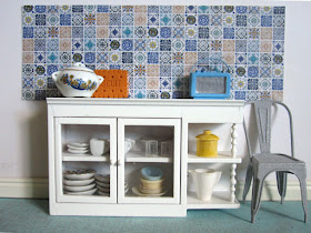 Modern one-twelfth scale miniature kitchen cabinet and cafe chair in front of a wall of random tiles in shades of blue.