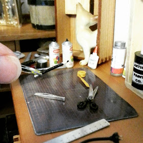 Hand using tweezers to place a on-twelfth scale pencil onto a miniature worktable top containing a cutting mat and various tools, including miniature tweezers.