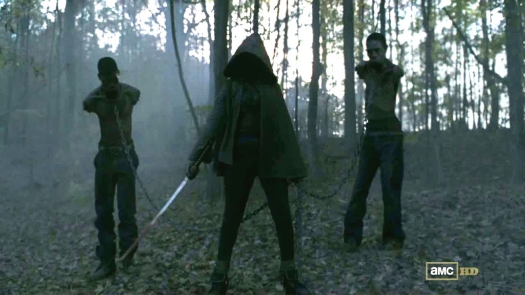 Michonne, cappuccio in testa e katana, porta con sé i due zombie incatenati, senza braccia e senza bocca
