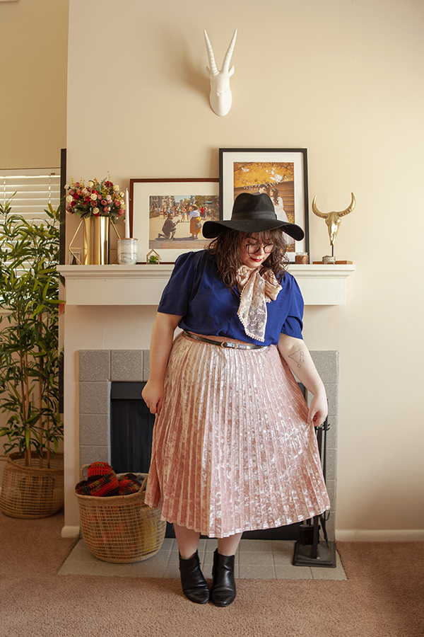An outfit of a large black wool floppy hat, short sleeved navy blue blouse, pastel pink midi skirt and a pink lace scarf tied into an off center bow around the neck.