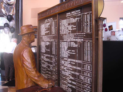 Sculpted figure of a man stands looking at a large metal sign listing train times