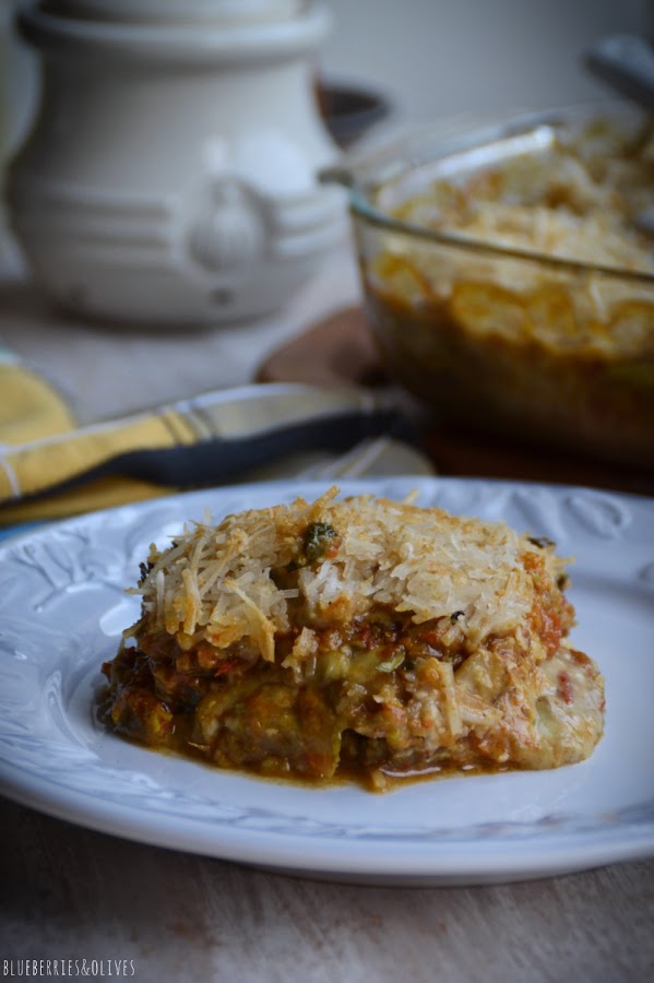Trozo de lasaña cortado sobre plato blanco, Lasaña de berenjenas en fuente de cristal sobre tabla de madera, con ingredientes de fondo, hierbas aromáticas, laurel seco, molinillo pimienta
