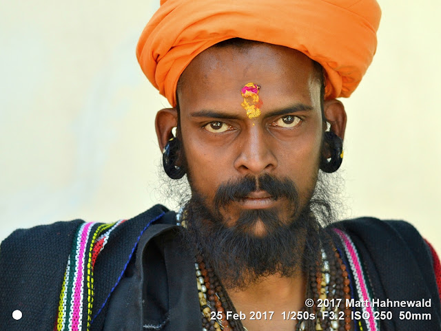 matt hahnewald photography; facing the world; gorakh nath; monk; monastery; bhavnath; bhavnath fair; character; face; earrings; ears; holed; eyes; facial expression; eye contact; full beard; turban; orange; consent; empathy; rapport; traveling; religious; traditional; cultural; hinduism; festival; event; mela; devotee; pilgrim; junagadh; gujarat; asian; indian; western india; one person; male; young; man; picture; photo; face perception; physiognomy; educational; nikon d3100; nikkor af-s 50mm f/1.8g; prime lens; 50mm lens; 4x3 aspect ratio; horizontal orientation; street; portrait; closeup; headshot; full-face view; outdoors; color; posing; authentic; kanphata; yogi; darshani; gorakhnathi; shaivism; tilaka; forehead; stern; grimly; splitted ears