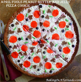 April Fools Peanut Butter and Jelly Pizza Cookie, a fun trick to play on the family. Peanut Butter Cookie, jelly in the frosting and lots of fun sweet toppings. | Recipe developed by www.BakingInATornado.com | #AprilFoolsDay #recipe #cookie