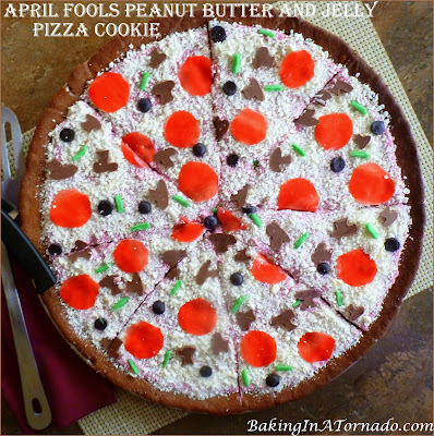 April Fools Peanut Butter and Jelly Pizza Cookie, a fun trick to play on the family. Peanut Butter Cookie, jelly in the frosting and lots of fun sweet toppings. | Recipe developed by www.BakingInATornado.com | #AprilFoolsDay #recipe #cookie