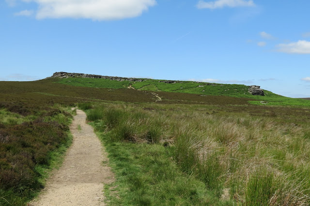 A sandy path runs through moorland to an escarpment running across the horizon.