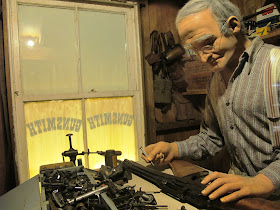 Full-scale model of the interior of an early 20th century gunsmith's shop, with a man working on a gun.