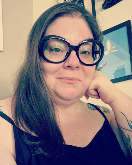image of me sitting at my desk wearing a black tanktop, with my hair down, and sporting giant black oval frame eyeglasses
