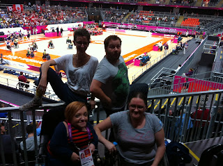 Picture of 4 people. The back row is a non-disabled woman and a non-disabled man perched on a railing. The front row is 2 women in wheelchairs. In the background wheelchair rugby players are on the court warming up.