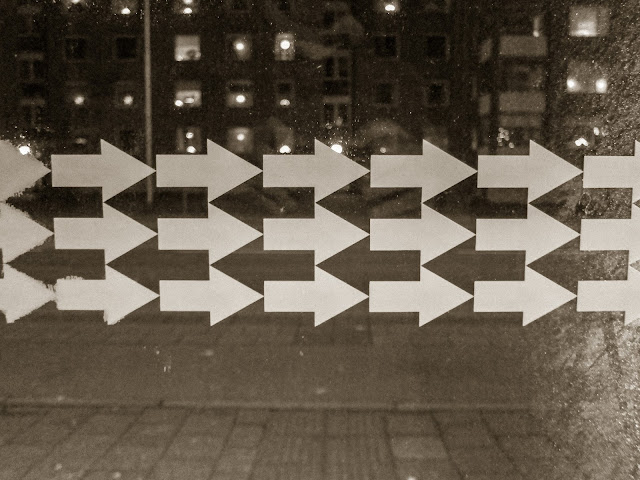Multitud de flechas blancas en tres líneas una encima de otra, fotografiadas sobre un panel de cristal a través del que se ven luces de farolas y edificios al fondo en una noche de ciudad.
