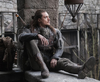 Uhtred de Bebbanburg sentado, apoyando la espalda en una columna. Tiene el pelo largo, viste con ropa de guerrero, y lleva a la espalda su famosa espada con un ámbar en la empuñadura.