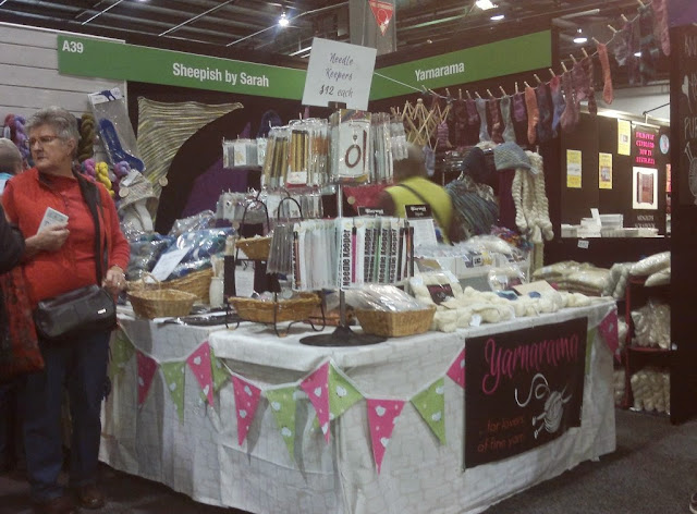 Sheepish by Sarah and Yarnarama combined stall has an L-shaped table at the corner which is decorated with pink and green bunting and a black Yarnarama sign. Undyed yarn is laid out on the table above the black sign with dye powders next to them. An assortment of accessories are crammed onto the left hand side of the table. Above, on the right one can see a row of socks hanging from a line. A knitted shawl can be seen on the black wall on the left.  Skeins of yarn are displayed along the wall behind the woman in the red top on the left hand side of the picture.