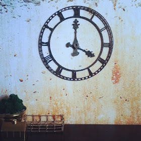Industrial miniature scene of a large metal clock on a distressed wall behind a desk with various brass items displayed on it.