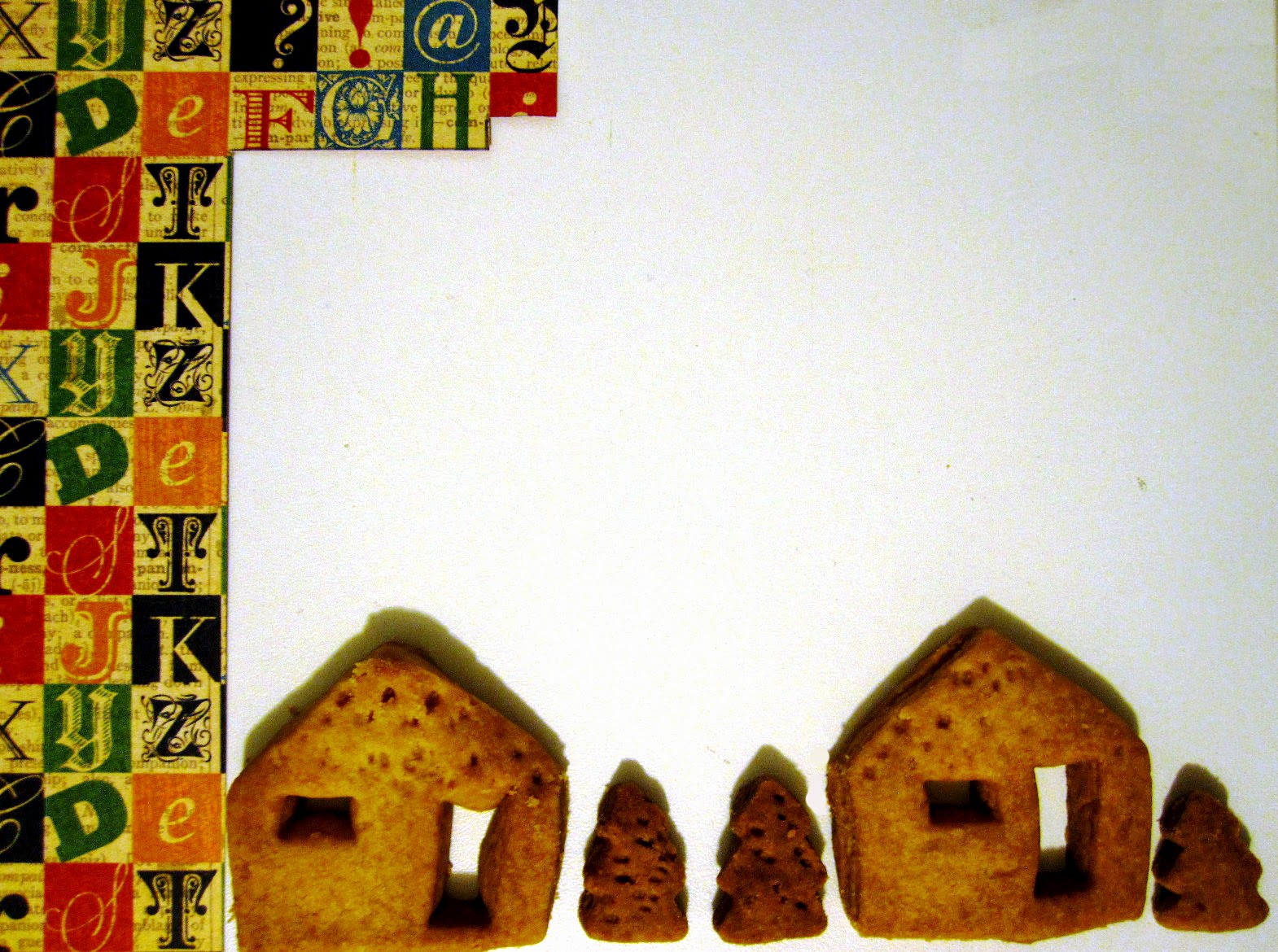 Part of a piece of scrapbooking paper with a typographic design next two several shortbread biscuits in the shape of houses and trees.
