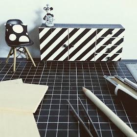 One-twelfth scale modern miniature sideboard with black and white striped front, next to an Eames chair with a black and white spotted cushion on it, on a cutting mat with various full-sized tools laid out on it.