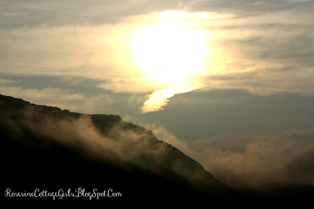 What does God Sound Like? Photo of the sunrise coming up over rough craggy mountains. The sun is brilliant in the sky with rays coming off of it and lighting the cloudy sky. The mountains are dark in the foreground and there are whisps of clouds hugging their peaks. by RosevineCottageGirls.com  