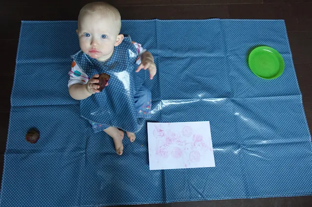 An over head photograph of a baby on a splash mat, wearing a tunic bib and holding a piece of cut beetroot. A white piece of card with pink marks is in front of her